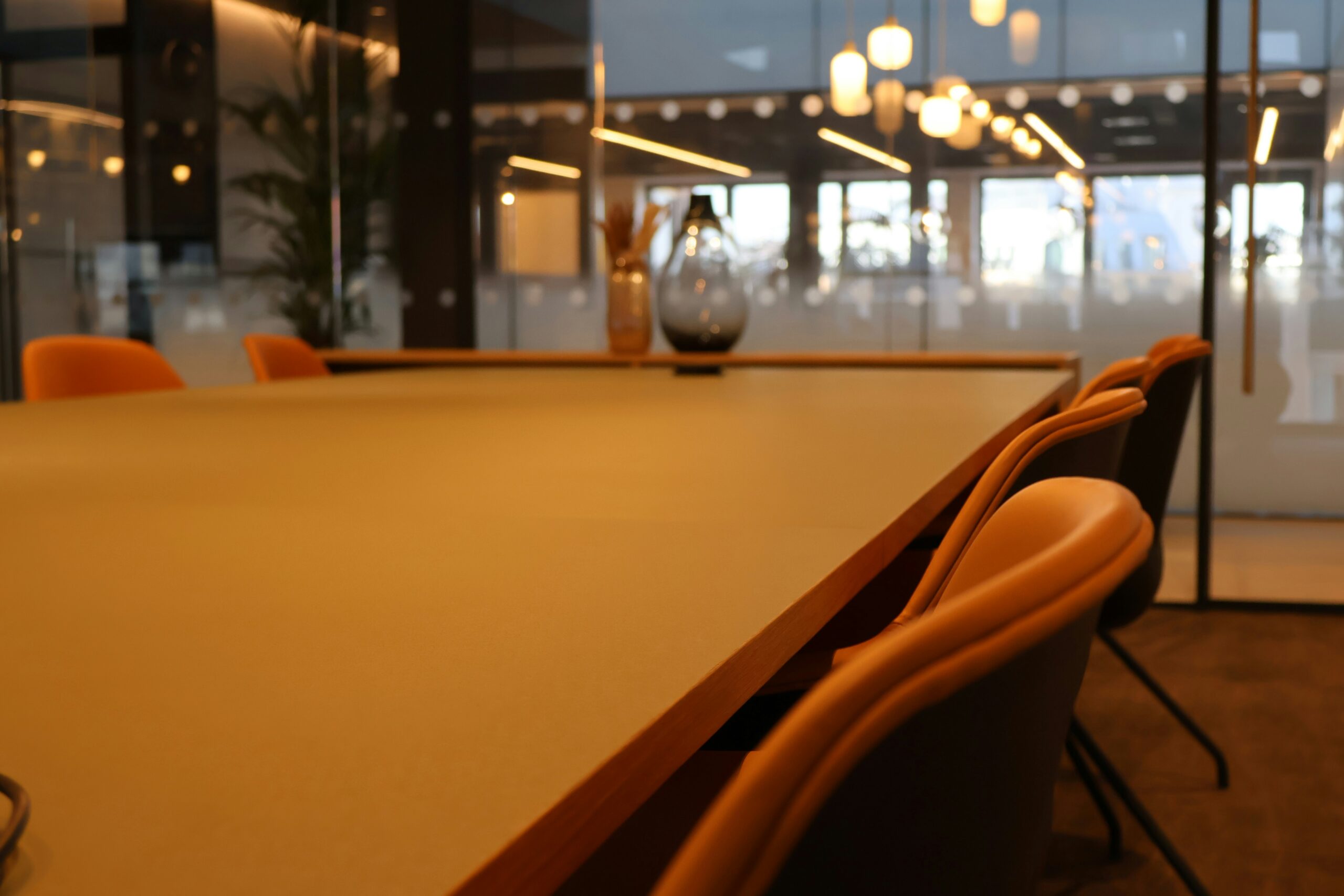A close-up, angled view of a long, modern wooden conference table illuminated by warm, ambient overhead lighting, featuring empty orange and black office chairs arranged along the side with a softly blurred background.